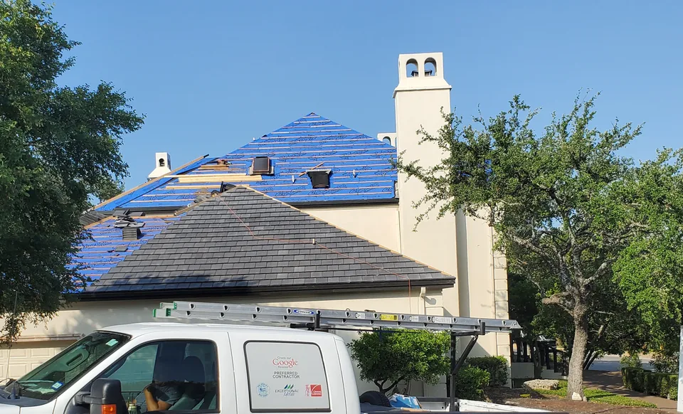Roof work underway on a tile roof: blue underlayment and wood battens on the upper slopes, finished tile on the lower roof.