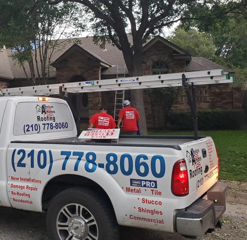 Two American Homes Roofing crew members at a ladder against a brick home with a shingle roof, with the company pickup parked in front.