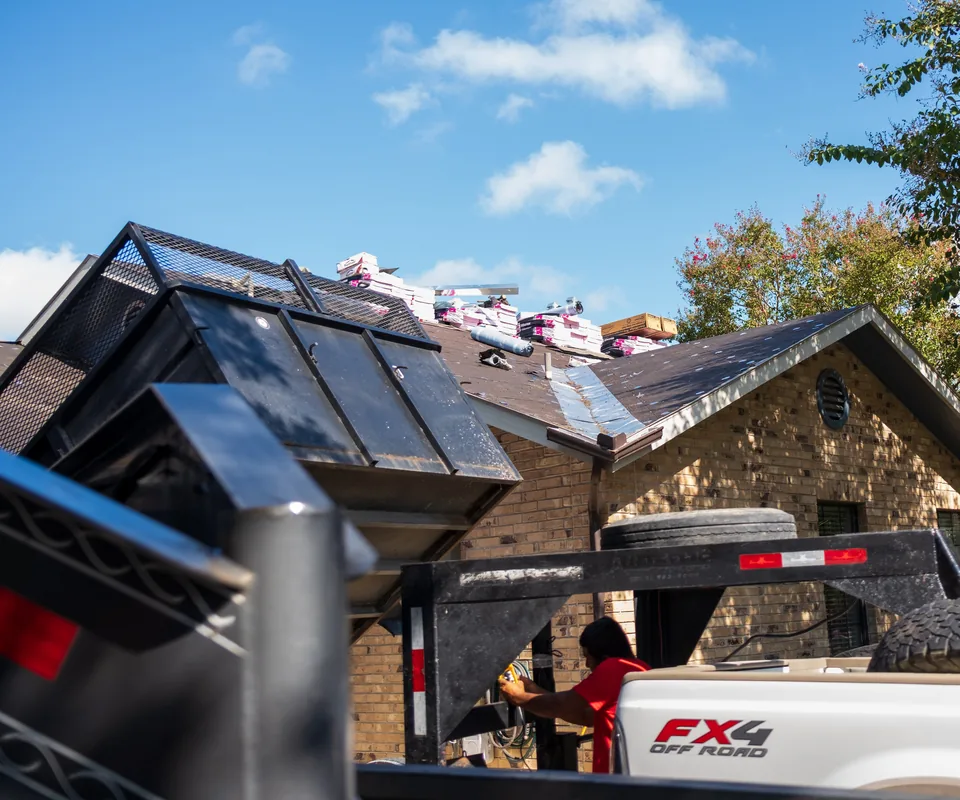 Bundles of new shingles staged on a roof during a replacement, seen past a dump trailer, with a crew member working beside the house.