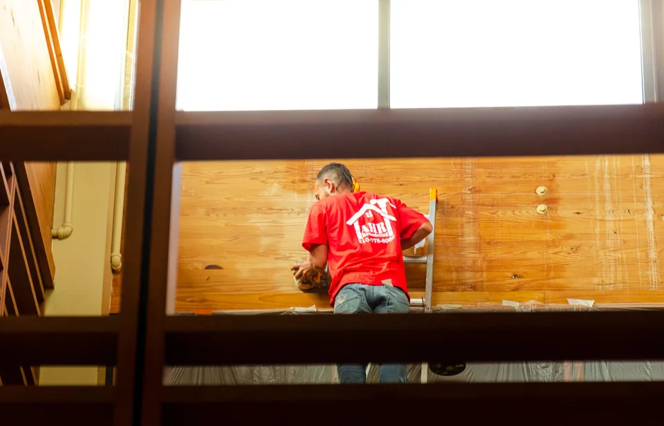 AHR crew member on a ladder sanding a high wood wall inside a commercial building, with the floor below covered in plastic.