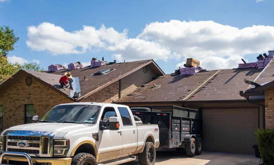 American Homes Roofing crew member on a roof being replaced, with bundles of new shingles along the ridge and a pickup and dump trailer in the driveway.