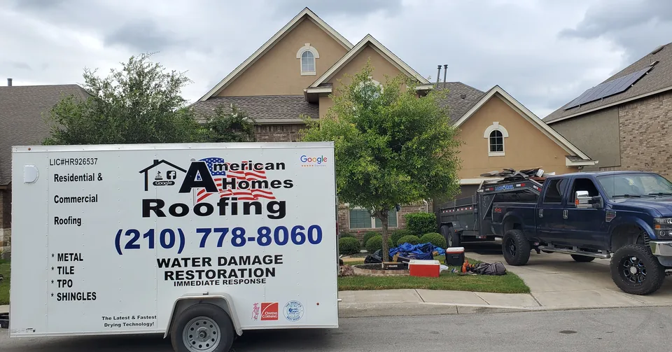 American Homes Roofing trailer parked in front of a two-story home, with a loaded dump trailer in the driveway.